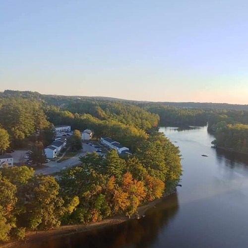 A serene river scene with autumn foliage, surrounded by trees and a few buildings along the shore under a clear sky.