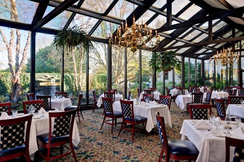 Bright restaurant interior with glass walls, elegant tables, and lush greenery outside. Cozy and inviting atmosphere.