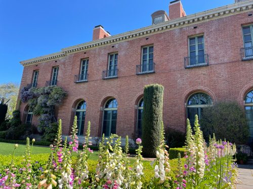 A brick building with arched windows, surrounded by lush greenery and colorful flowers under a clear blue sky.