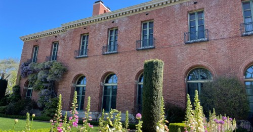 A brick building with large windows, surrounded by lush greenery and colorful flowers under a clear blue sky.