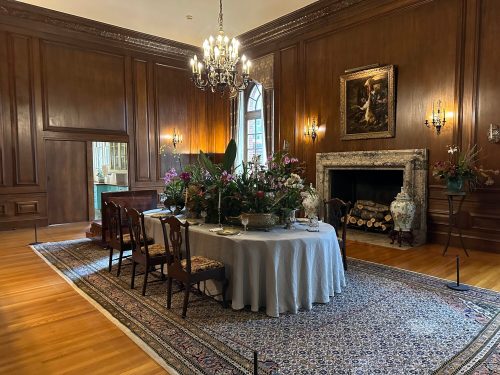 A beautifully arranged dining room with a large table, floral centerpieces, wooden walls, and a cozy fireplace.