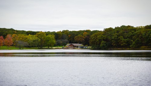A serene lake surrounded by trees in autumn colors, with a rustic building visible on the shore.