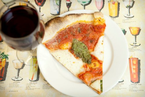 A slice of pizza with tomato sauce and basil on a plate, next to a glass of red wine, on a colorful drink menu background.