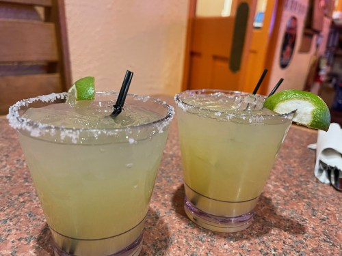 Two margaritas with salt rims and lime wedges, served in clear glasses on a textured countertop.