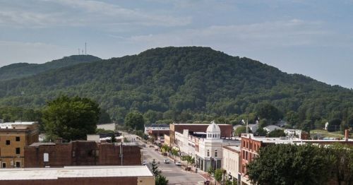 A view of a small town with historic buildings, trees, and a large green hill in the background under a clear sky.