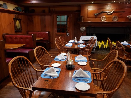 Cozy restaurant interior with wooden tables, chairs, and a fireplace, set for dining with blue napkins and white plates.