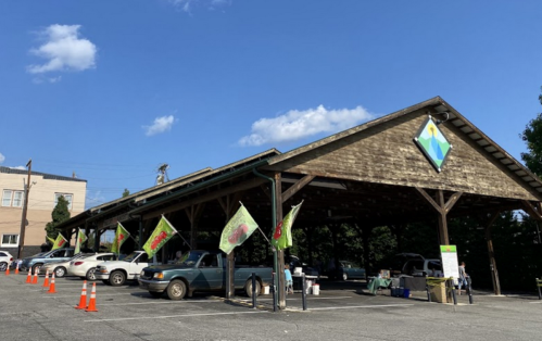 A covered market with cars parked outside, featuring colorful flags and a clear blue sky.