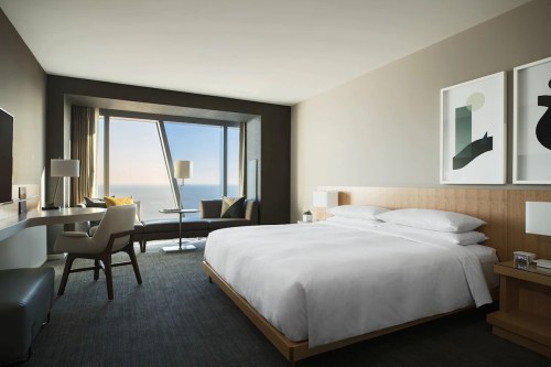 Modern hotel room with a large bed, desk, and a window showcasing a scenic ocean view. Minimalist decor.