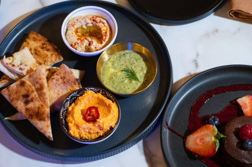 A black plate with hummus, green dip, pita bread, and a dessert featuring berries and chocolate.