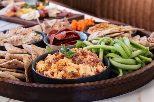 A wooden platter filled with various snacks: dips, crackers, celery, pepperoni, and vegetables.
