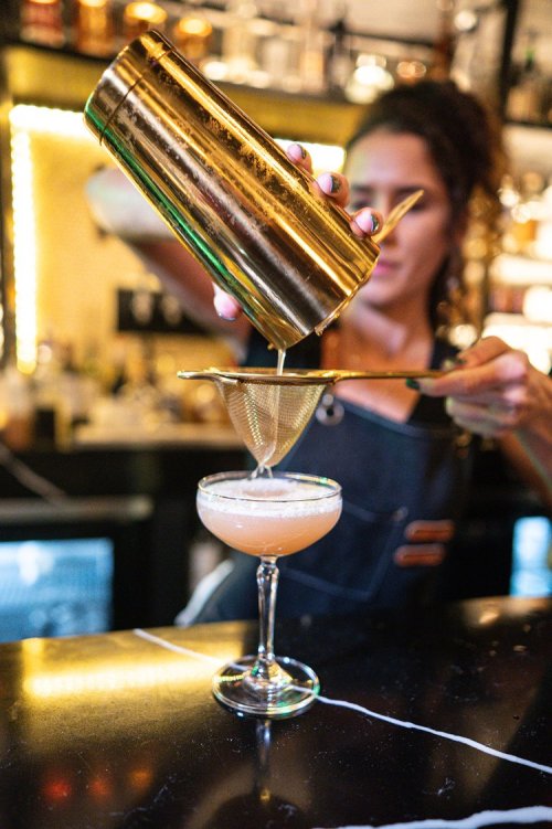 A bartender pours a cocktail from a shaker into a glass, using a strainer to catch any solids.