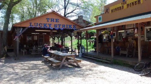A rustic outdoor dining area with picnic tables, featuring two buildings: "The Lucky Strike" and "The Miners Diner."