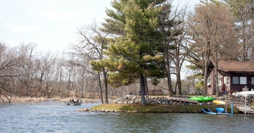 Brown lakeside cabin with kayaks on the shore and a fishing boat on a quiet Minnesota lake.