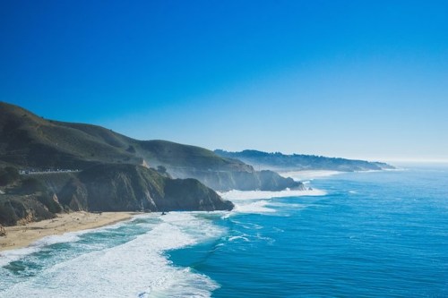 A scenic coastal view with waves crashing on the shore, surrounded by hills under a clear blue sky.