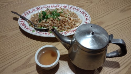 A plate of fried rice topped with green onions, accompanied by a silver teapot and a small bowl of tea.