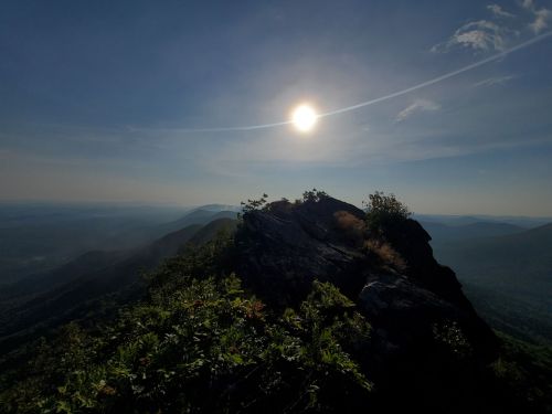 A sunlit mountain peak with lush greenery, overlooking a vast valley under a clear blue sky.