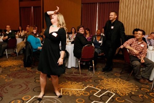 A woman in a black dress playfully poses while others watch at a lively event with tables and chairs in the background.