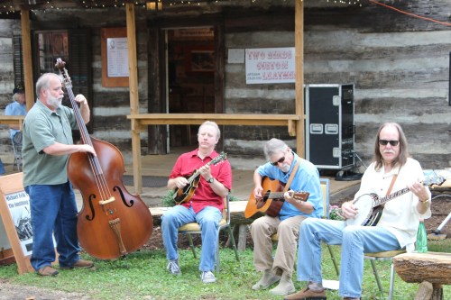 Four musicians play instruments outdoors near a rustic wooden building, creating a lively atmosphere.