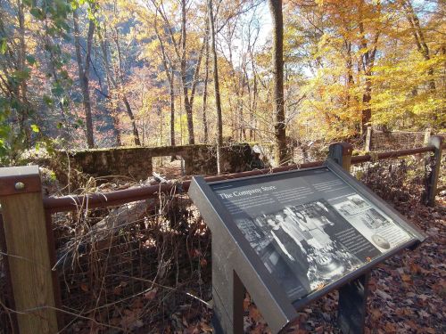 A historical sign in a forest with autumn foliage, near the ruins of a stone structure.