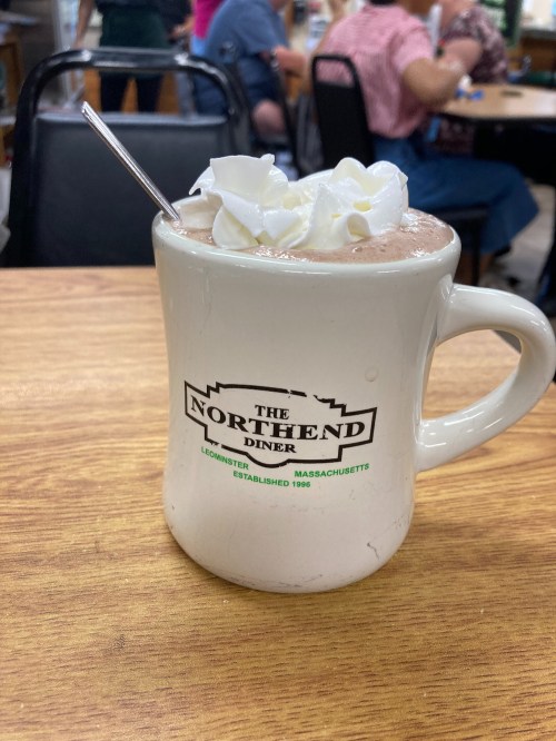 A mug from The North End Diner filled with hot chocolate topped with whipped cream, placed on a wooden table.