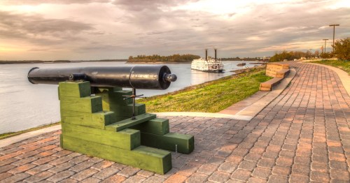 A view of the Mississippi River from atop of the levee at New Madrid, Missouri. A black metal cannon rests atop pavers in the foreground, and a white steamboat is docked downriver.