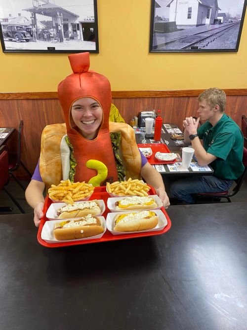 A person in a hot dog costume holds a tray with hot dogs and fries in a diner setting.