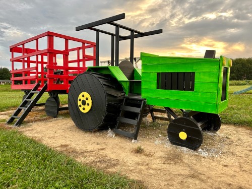 Colorful wooden tractor and wagon play structure in a grassy field under a cloudy sky.