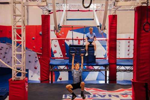 A child hangs from gymnastic rings while another sits on a platform in a colorful indoor obstacle course.