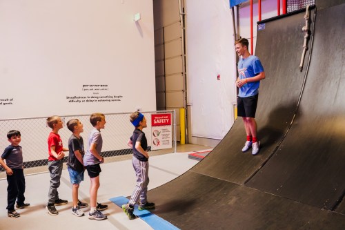 A group of children watches as a young man stands on a ramp, demonstrating a trick at a skate park.