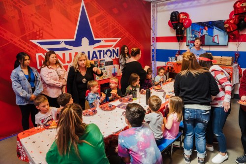 A lively birthday party scene with children and adults gathered around a decorated table, celebrating in a colorful venue.