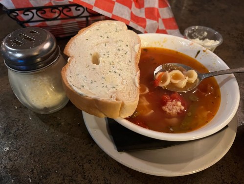 A bowl of soup with pasta and vegetables, accompanied by a slice of garlic bread and a container of grated cheese.