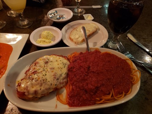 A plate of chicken parmesan with melted cheese, spaghetti in marinara sauce, butter, bread, and a drink on the side.