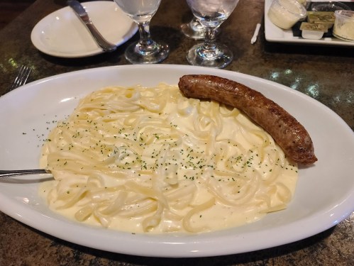A plate of fettuccine alfredo topped with a grilled sausage, garnished with parsley, on a restaurant table.