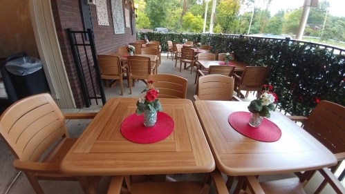Outdoor dining area with wooden tables, chairs, and potted flowers on red placemats, surrounded by greenery.