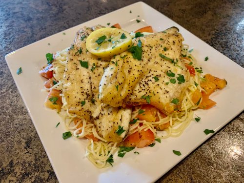 A plate of grilled fish fillets on a bed of pasta and diced tomatoes, garnished with a lemon slice and parsley.