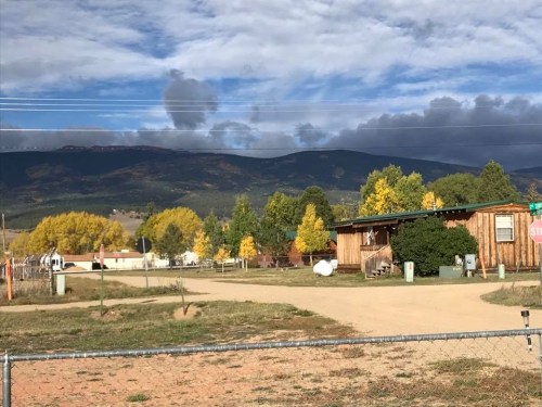 A rural scene featuring a wooden house, colorful trees, and mountains under a cloudy sky.