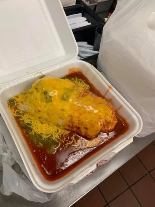 A takeout container with a large burrito smothered in cheese and sauce, surrounded by green peppers.