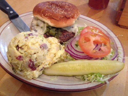 A plate with a sesame seed burger, potato salad, lettuce, tomato, onion, and a pickle on the side.