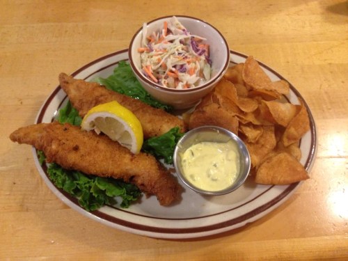 A plate with two fried fish fillets, a lemon wedge, coleslaw, chips, and a small bowl of dipping sauce.