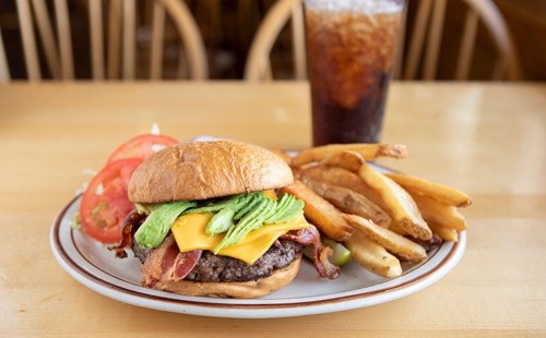 A juicy burger with bacon, cheese, and avocado, served with fries and a glass of cola on a wooden table.
