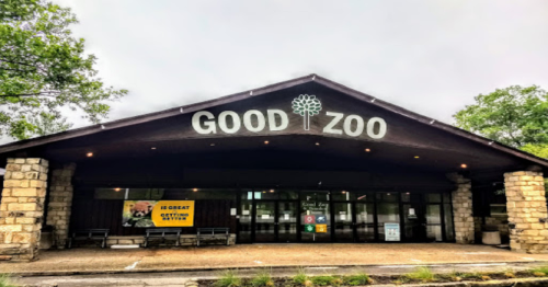 Entrance of Good Zoo with a stone facade and large sign, surrounded by trees under a cloudy sky.