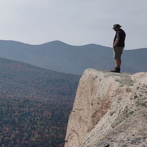 A person stands on the edge of a rocky cliff, overlooking a vast landscape of mountains and colorful trees.