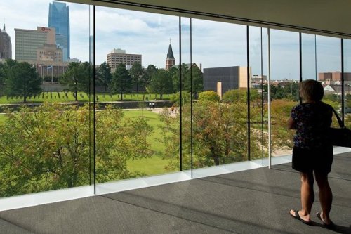 A person stands by a large window, overlooking a cityscape with trees and buildings in the background.