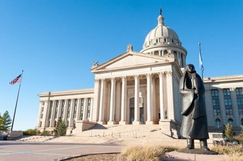 A statue of a woman stands in front of a grand government building with a dome, under a clear blue sky.