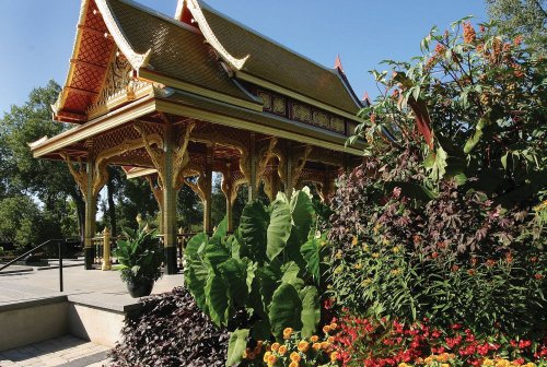 A vibrant garden with colorful flowers surrounds a traditional Thai pavilion under a clear blue sky.