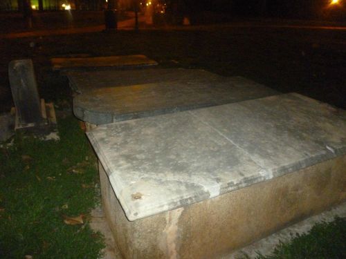 A weathered stone tomb or grave marker in a dimly lit park, surrounded by grass and trees.