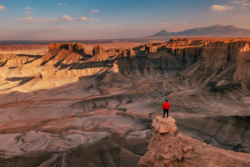 A person in a red jacket stands on a rocky outcrop, overlooking a vast, rugged desert landscape at sunset.