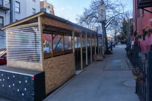 A wooden outdoor dining structure with clear plastic walls, set along a city sidewalk on a sunny day.