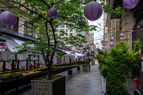 A street scene with purple lanterns hanging above outdoor seating areas, surrounded by greenery and urban buildings.