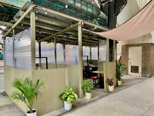 Outdoor dining area with covered booths, plants, and string lights, located near an elevated train track.